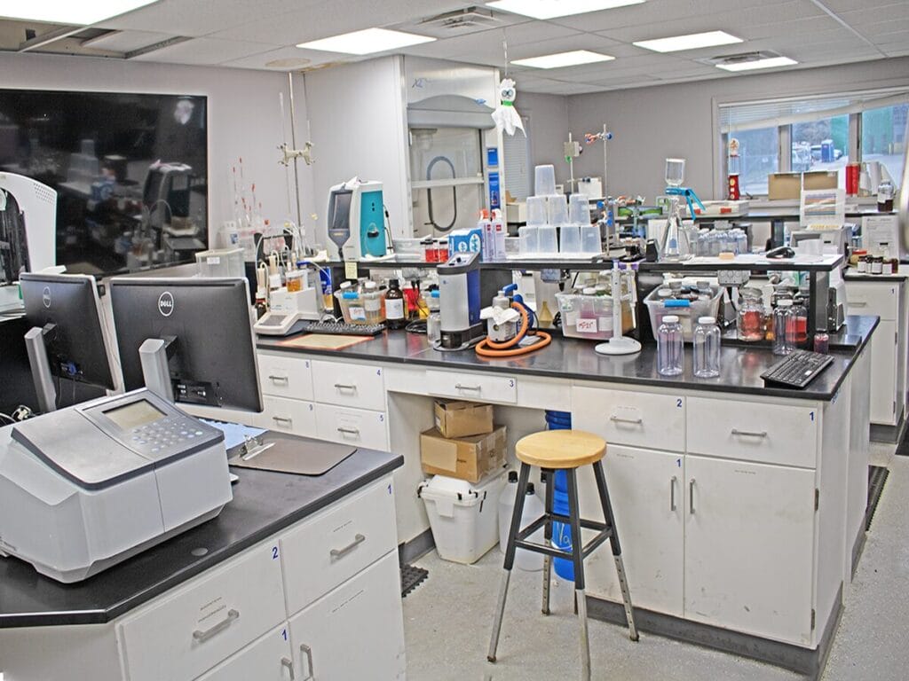 The interior of a fuel testing laboratory with workbenches equipped with analytical instruments, computers, glassware, and chemical samples used for fuel quality analysis and testing.