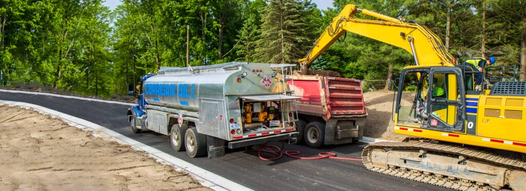 A Corrigan Oil Co. polished silver fuel and lubricant tanker truck parked on a freshly paved road at a wooded construction site, with a yellow excavator and red dump truck visible nearby.