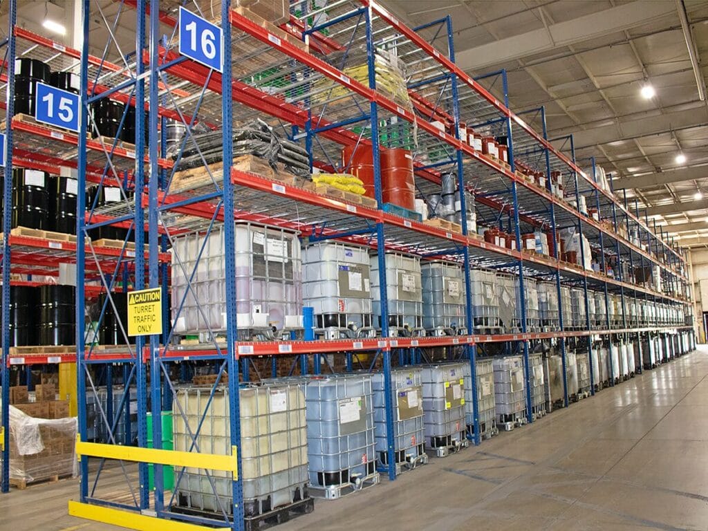 A large industrial warehouse with tall blue and red shelving racks stocked with bulk IBC tote containers, drums, and palletized lubricant and oil products, with a caution sign indicating turret traffic only.