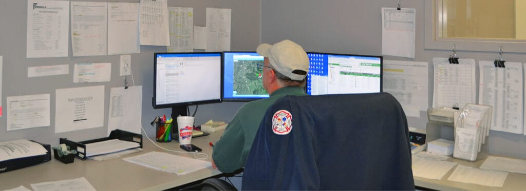 A Corrigan Oil Co. fuel dispatch coordinator viewed from behind, monitoring three screens displaying mapping software, delivery schedules, and fleet tracking data, with printed route sheets and delivery notes covering the surrounding walls.