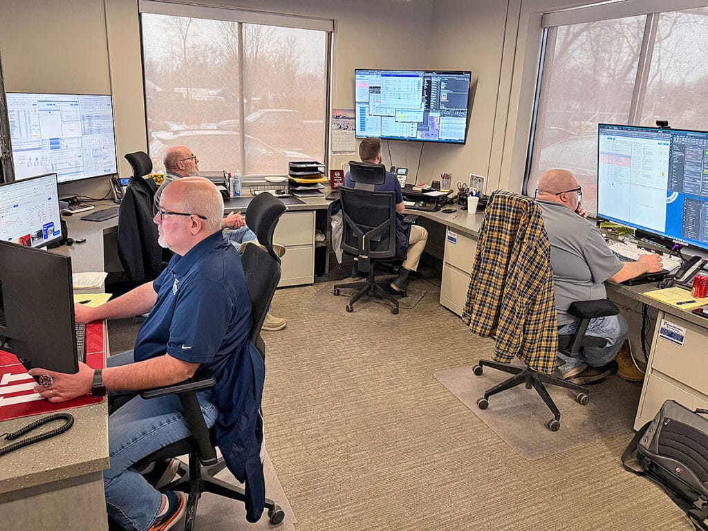 A fuel dispatch operations center with four employees working at computer workstations surrounded by multiple monitors displaying dispatch and logistics software.