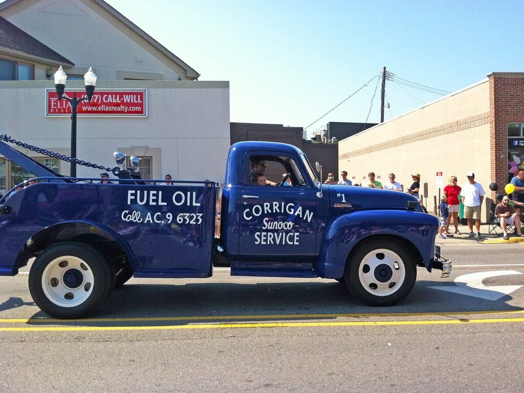 A restored vintage blue Chevrolet Corrigan Sunoco Service fuel oil delivery truck participating in a community parade, with spectators watching from the sidewalk in a downtown setting.