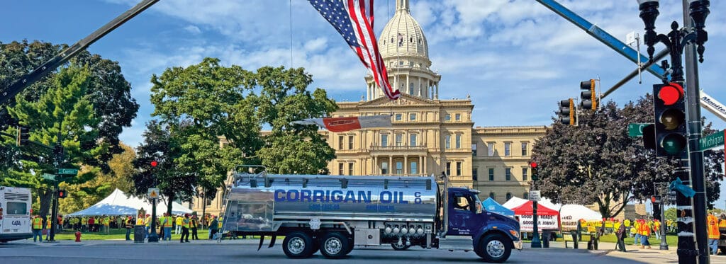 A Corrigan Oil Co. polished silver tanker truck parked on a street intersection in front of the Michigan State Capitol building during an outdoor public event, with a large American flag suspended overhead and workers in high-visibility vests gathered on the lawn.