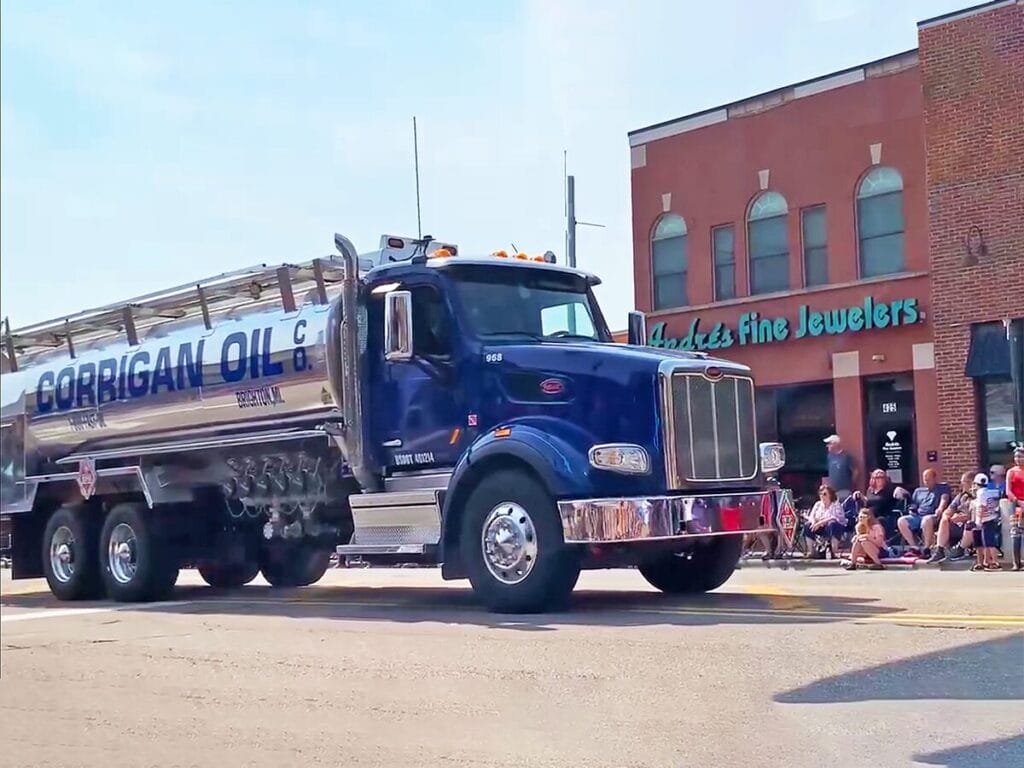A Corrigan Oil Co. blue Peterbilt tanker truck with a polished silver tank participating in a downtown community parade, passing in front of a brick building and spectators lining the street.
