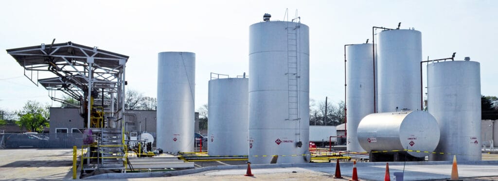 A commercial fuel storage facility with multiple large white vertical cylindrical tanks and horizontal tanks marked with combustible and flammable hazmat placards, surrounded by yellow safety barriers and traffic cones.