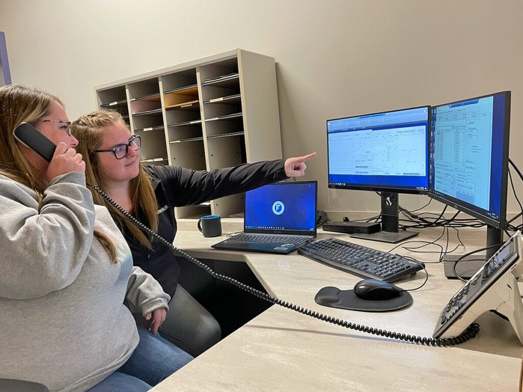 Two Corrigan Oil Co. customer service representatives working together at a desk, with one on the phone and the other pointing at information displayed across dual computer monitors