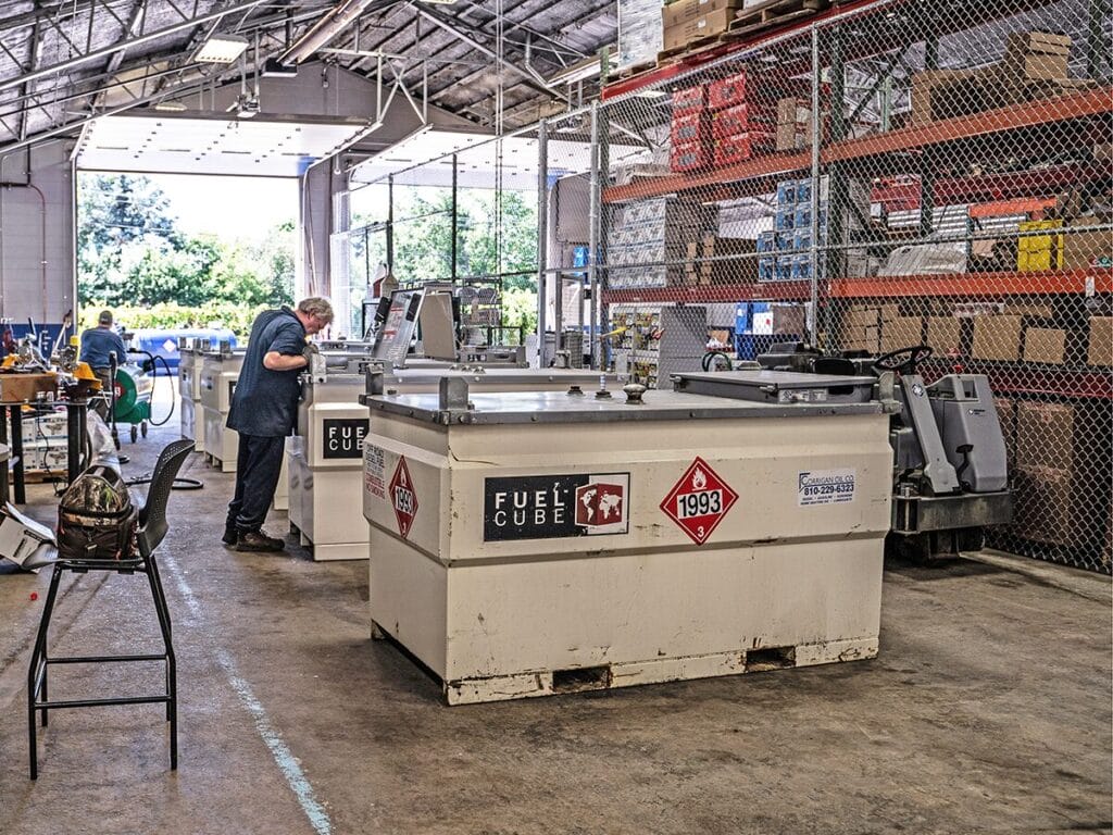 A technician inspects a row of white Fuel Cube portable diesel fuel storage units branded with Corrigan Oil Co. contact information inside an open warehouse facility.