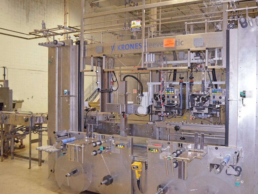 A large Krones Sleevematic industrial labeling and packaging machine on a production floor, used for high-speed automated sleeve label application in a manufacturing facility.