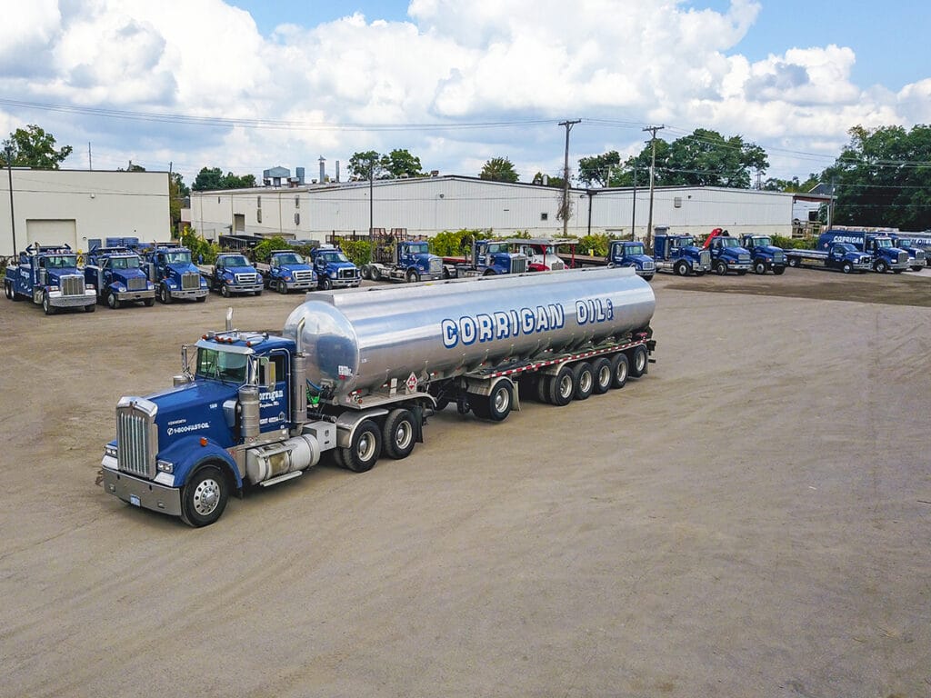 An aerial view of a large Corrigan Oil Co. Kenworth tanker truck with a polished silver tank in the foreground, with the company's full fleet of blue delivery trucks lined up outside their warehouse facility in the background.