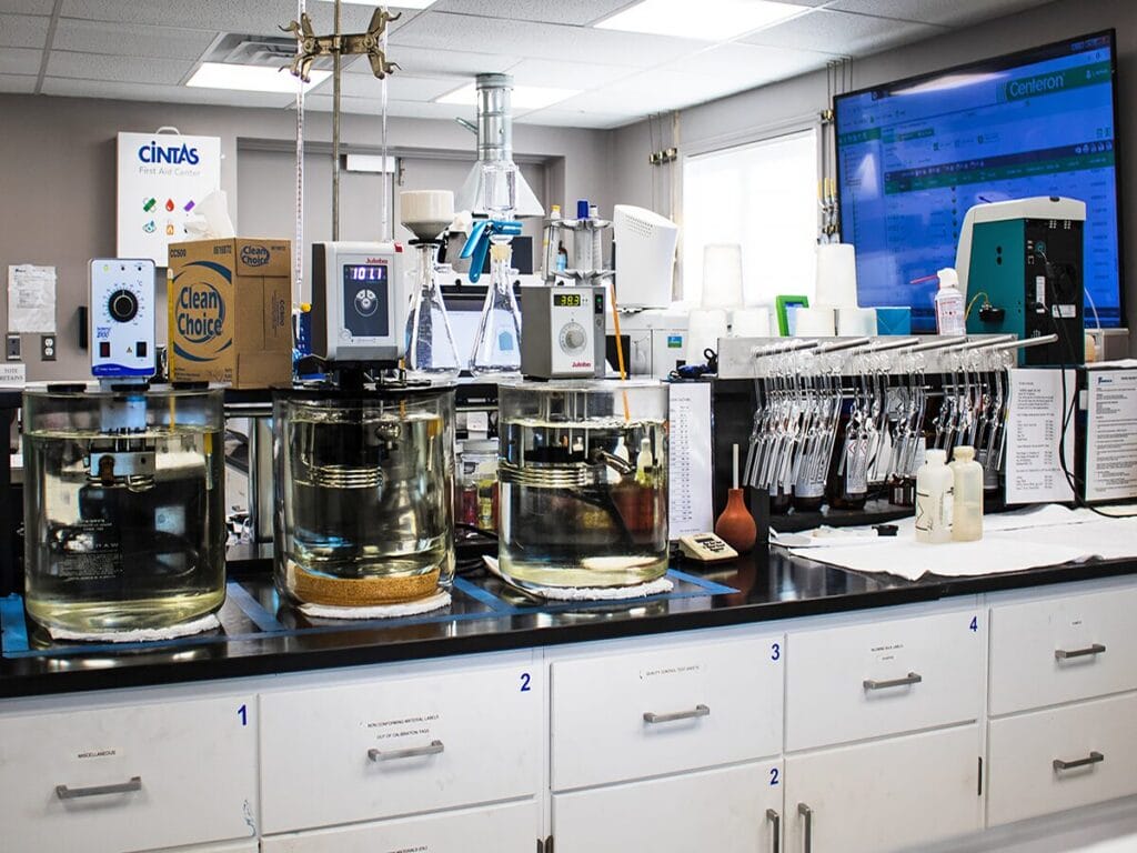A fuel testing laboratory workbench with large glass beakers containing fluid samples, a Julabo temperature control unit, rows of test tubes on a rack, and a monitor displaying the Centeron laboratory management system.