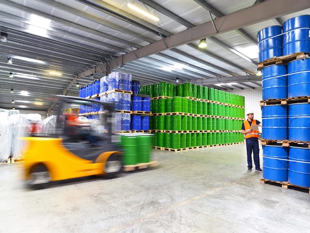 A warehouse worker in a high-visibility vest and hard hat reviews inventory on a clipboard while a yellow forklift moves green industrial drums, with large palletized stacks of green and blue barrels filling the facility.