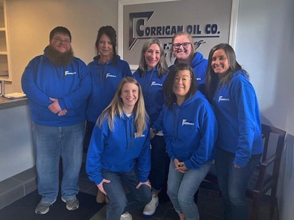 Seven Corrigan Oil Co. customer service team members posing together in matching blue branded hoodies in front of the company logo on the office wall.