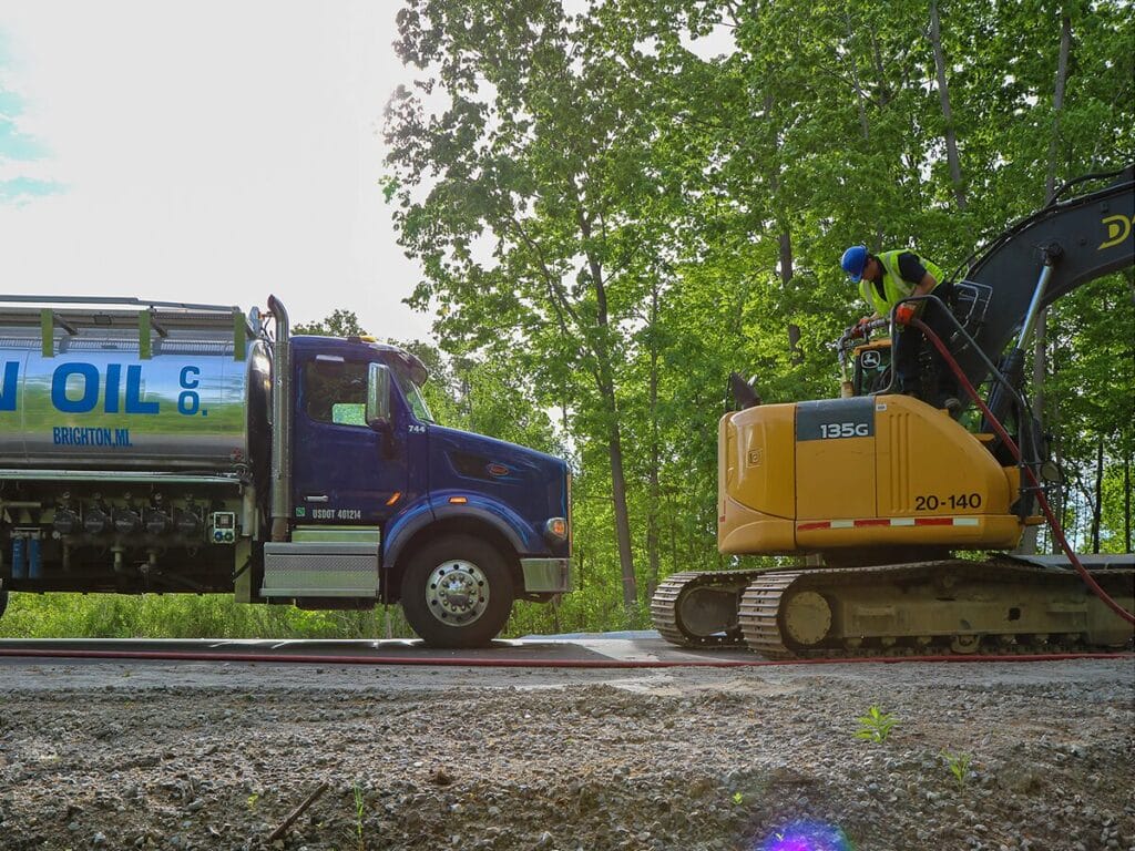 A female technician wearing safety glasses and black gloves applies Castrol Fifth Wheel Grease from a tube to a fifth wheel coupling on a commercial truck.