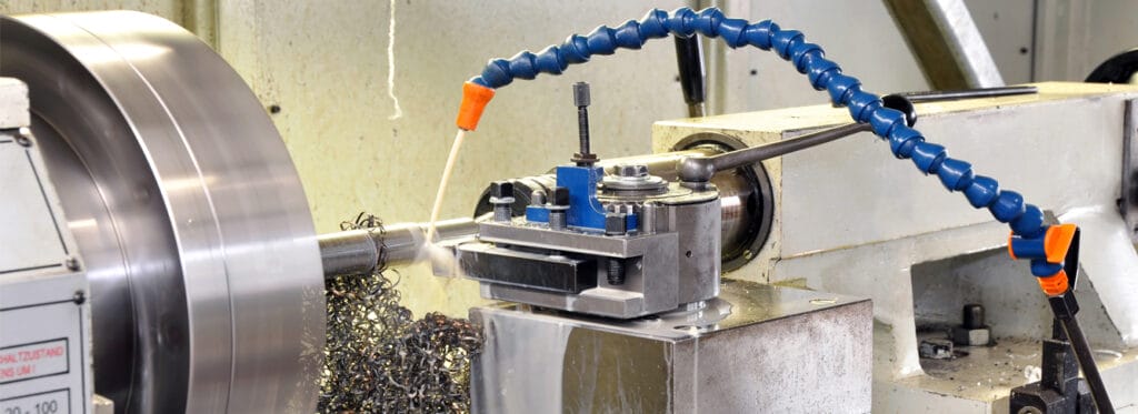 A metal lathe in operation with a flexible blue coolant hose delivering cutting fluid to the tool and workpiece, with metal shavings accumulated below the cutting area.