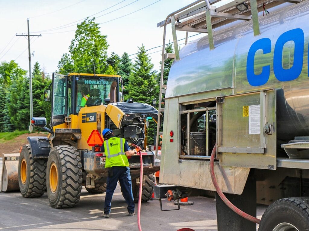 A Corrigan Oil Co. technician in a high-visibility vest and blue hard hat uses a red hose to service a yellow Komatsu WA270 wheel loader at an outdoor construction site.