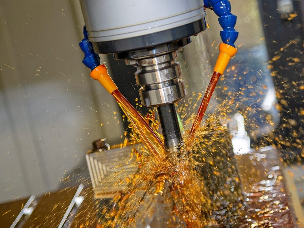 A close-up of a CNC milling machine spindle with cutting fluid streaming from orange nozzles onto the cutting tool, with metal chips and fluid splashing across the work surface.