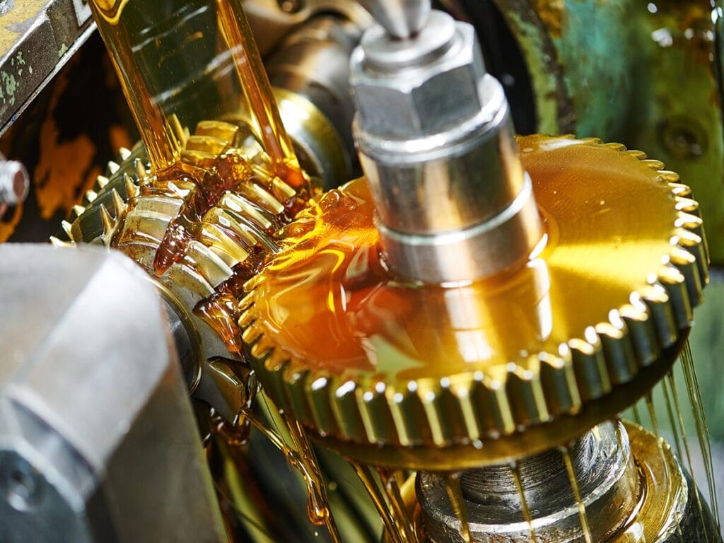 A close-up of golden industrial gear oil flowing over interlocking metal gears and a shaft in a manufacturing machine, with the oil coating the teeth of the gears.