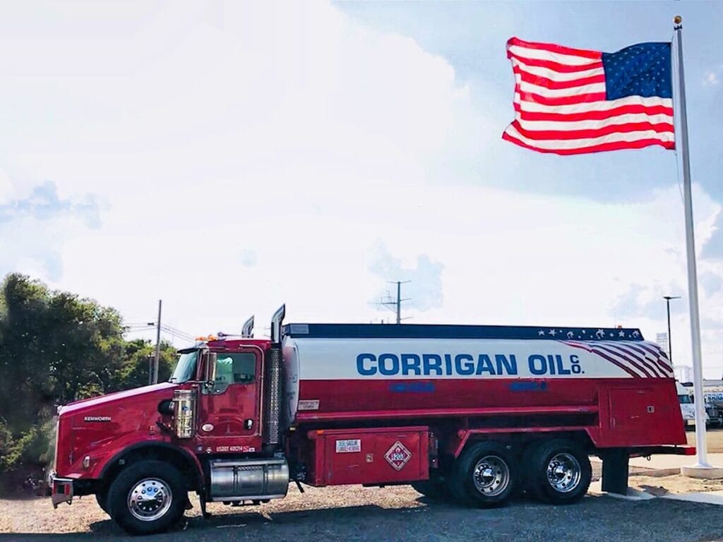 A red Kenworth Corrigan Oil Co. tanker truck with a patriotic stars-and-stripes themed tank parked beside a large American flag flying on a flagpole against a partly cloudy sky.