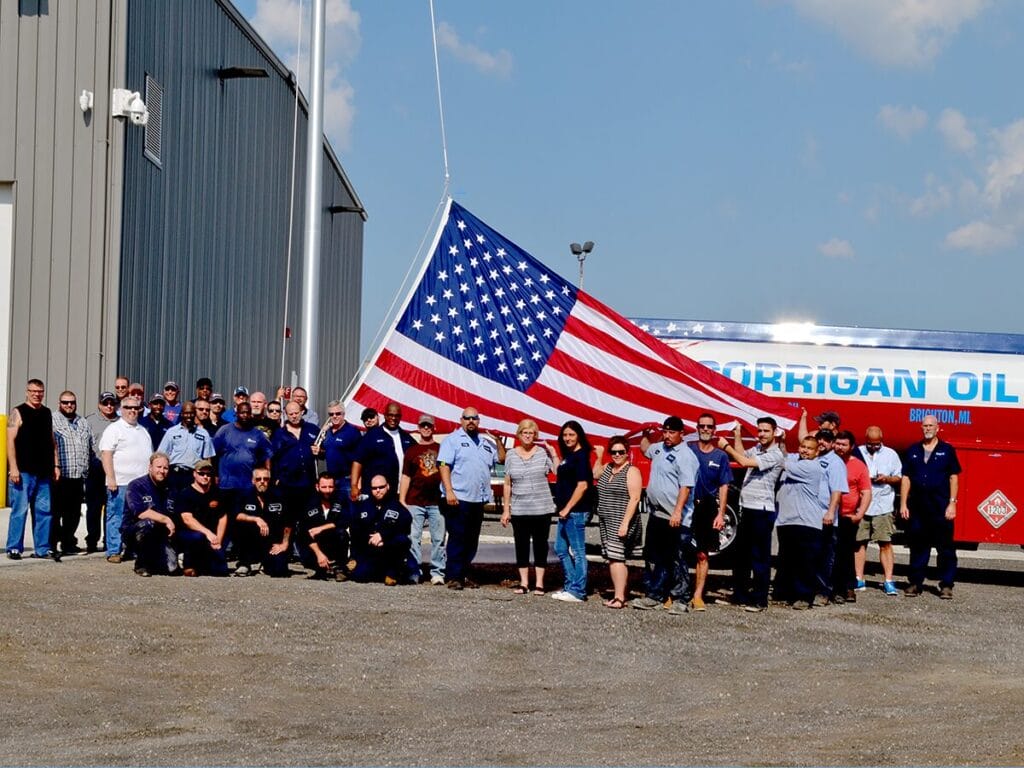 A large group of Corrigan Oil Co. employees posing together outdoors while holding an oversized American flag in front of the company's Brighton, MI facility and a patriotic red, white, and blue tanker truck.