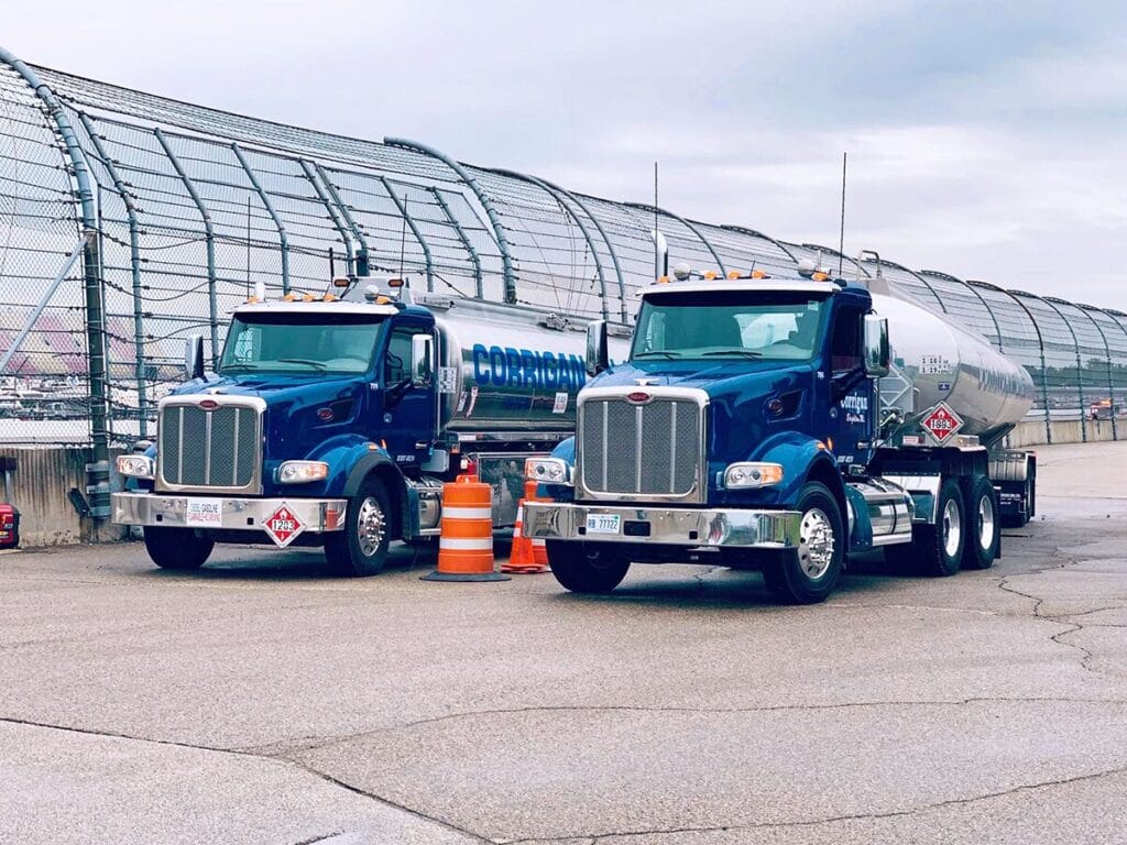 Two Corrigan Oil Co. blue Peterbilt tanker trucks with polished silver tanks parked side by side along the retaining wall of a motorsports racetrack, with orange safety cones between them.