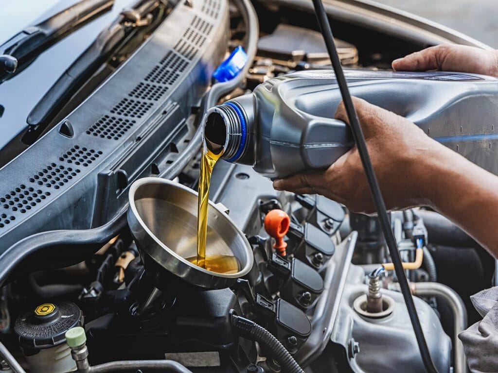 A close-up of a mechanic's hands pouring golden motor oil from a gray jug through a funnel into a car engine during an oil change