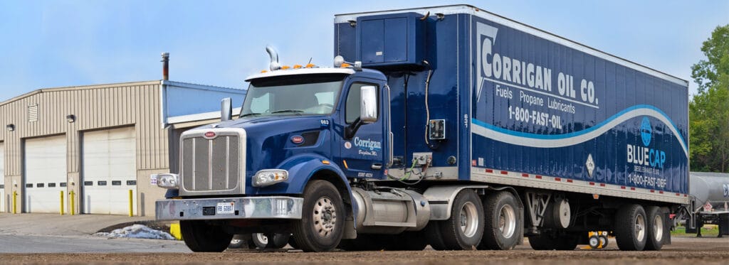 A Corrigan Oil Co. blue Peterbilt semi-truck with a branded trailer advertising fuels, propane, lubricants, and BlueCap diesel exhaust fluid, parked outside a commercial warehouse facility
