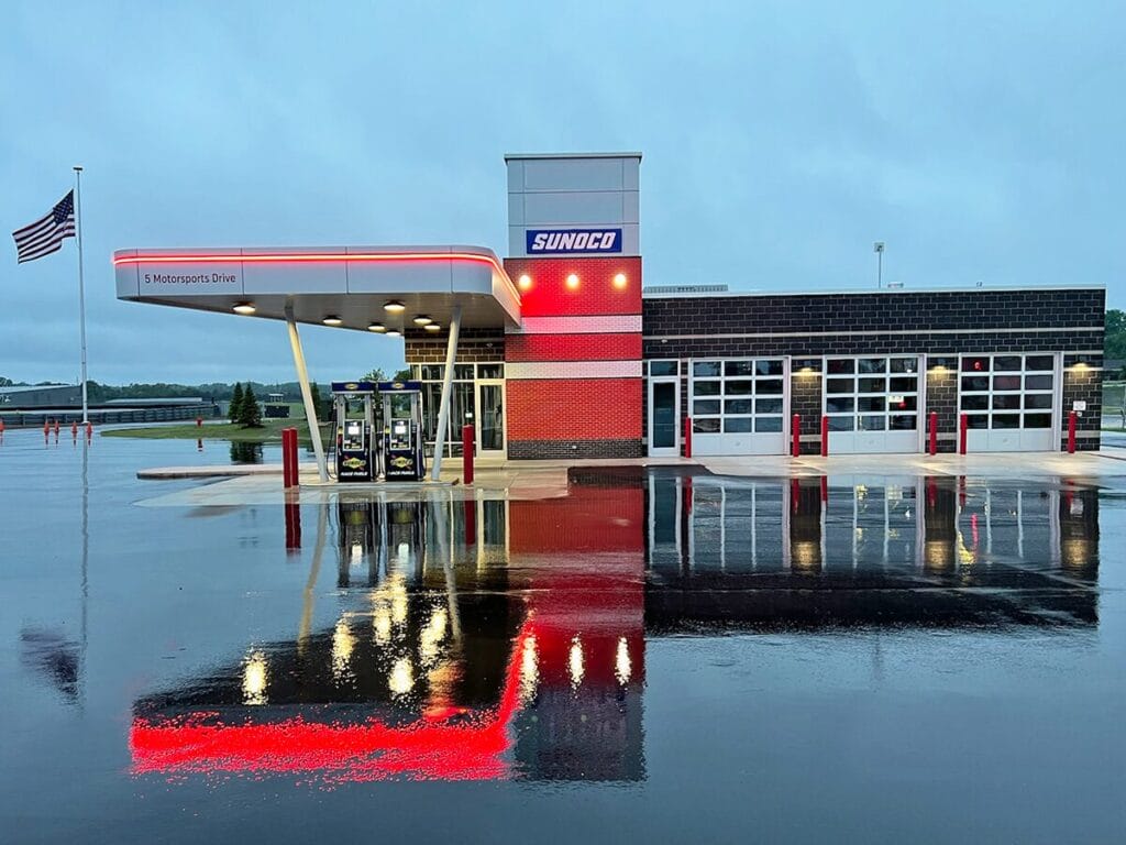 A modern Sunoco branded gas station and quick lube facility at dusk on a rainy evening, with the building and canopy lights reflecting in the wet pavement, located at 5 Motorsports Drive.